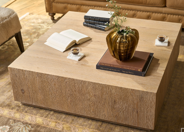 A square cocktail table with a light wood finish, featuring hidden casters, a vase, a book, and a cup on its surface, set in a living room setting.