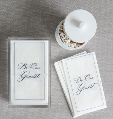 A set of white cloth napkins with the text 'BE OUR GUEST' printed in black, displayed in a clear plastic case alongside a small white dish with decorative white stuffing balls.