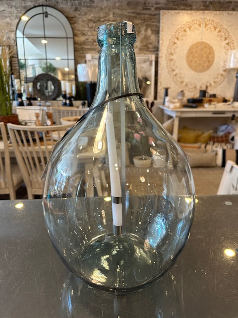 A large clear glass bottle with a swirling design, serving as a candle holder, displayed on a reflective surface with a white candle inside.