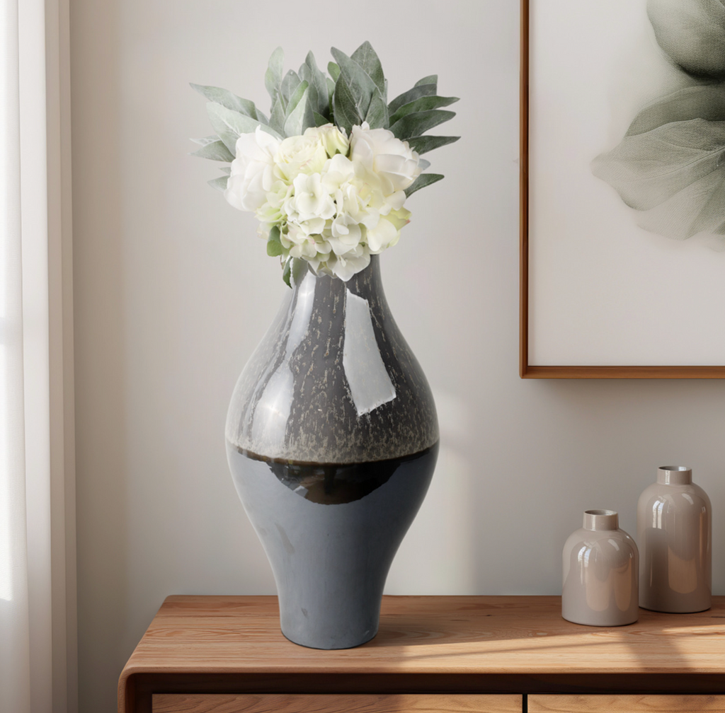 Gray textured, reflective vase with white flowers on a wooden table, framed artwork in the background