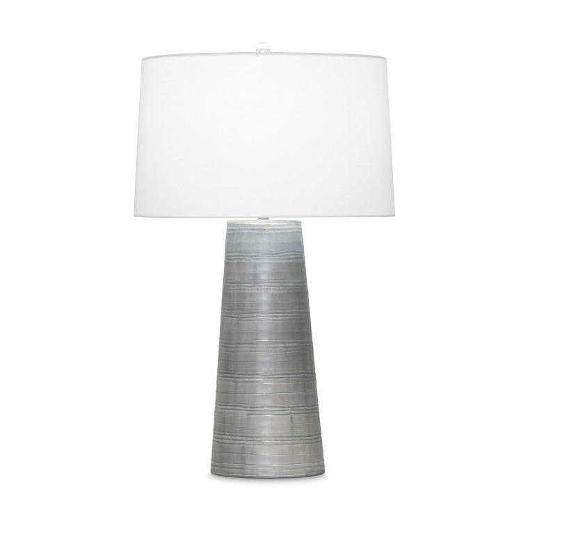 A table lamp with a tapered mouth-blown glass body in a grey carved finish, topped with an off-white linen drum shade.