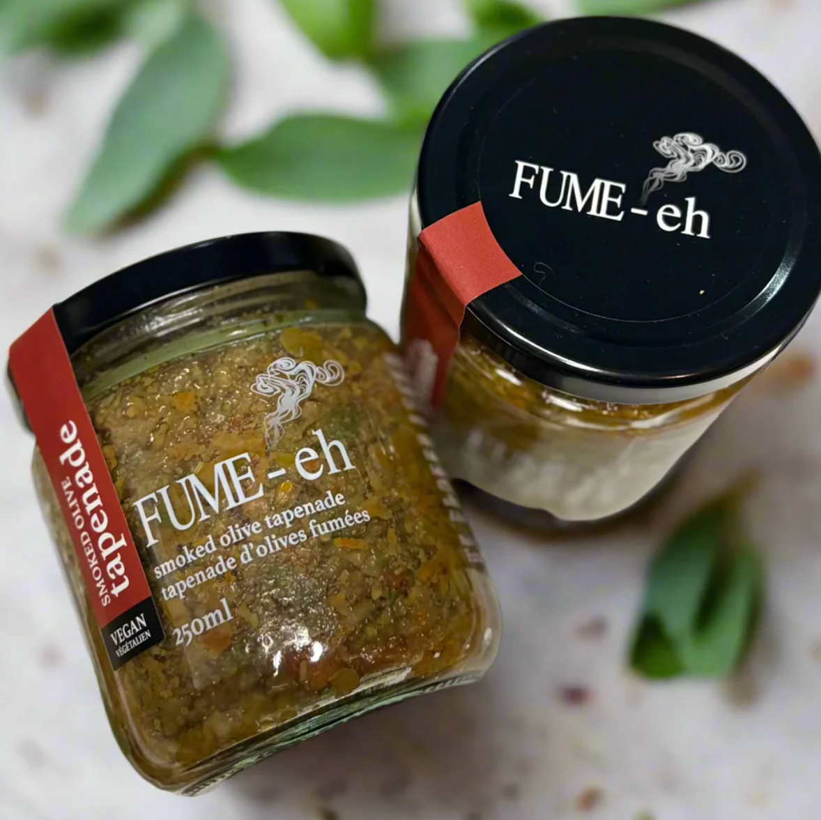 Two jars of FUME-eh smoked olive tapenade on a light background with green leaves.