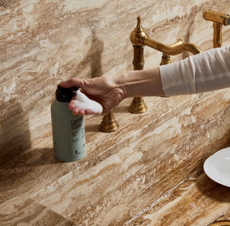 Person applying soap to a bar of soap on a marble countertop with gold fixtures.