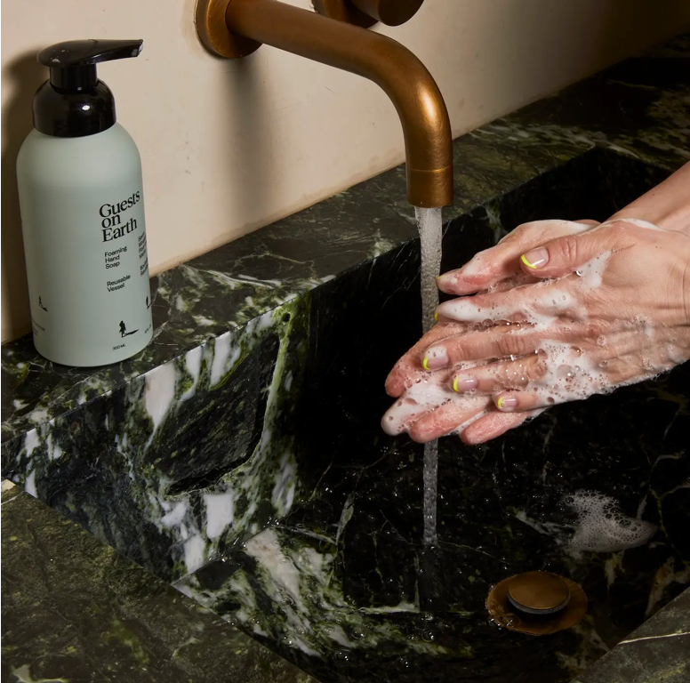 Person washing hands with soap under a running faucet on a marble sink, with a bottle of 'Gentle on Earth' soap visible.