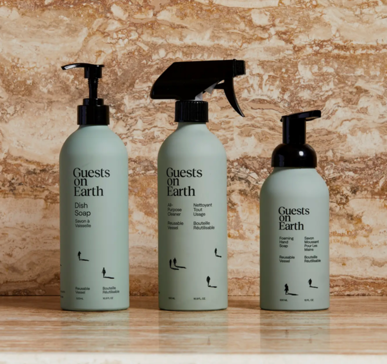 Three bottles of 'Guests on Earth' products on a wooden surface with a stone wall background.