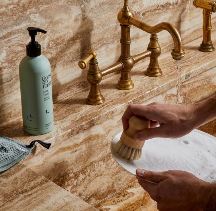Person cleaning a sink with a brush, next to a bottle of soap on a marble countertop.