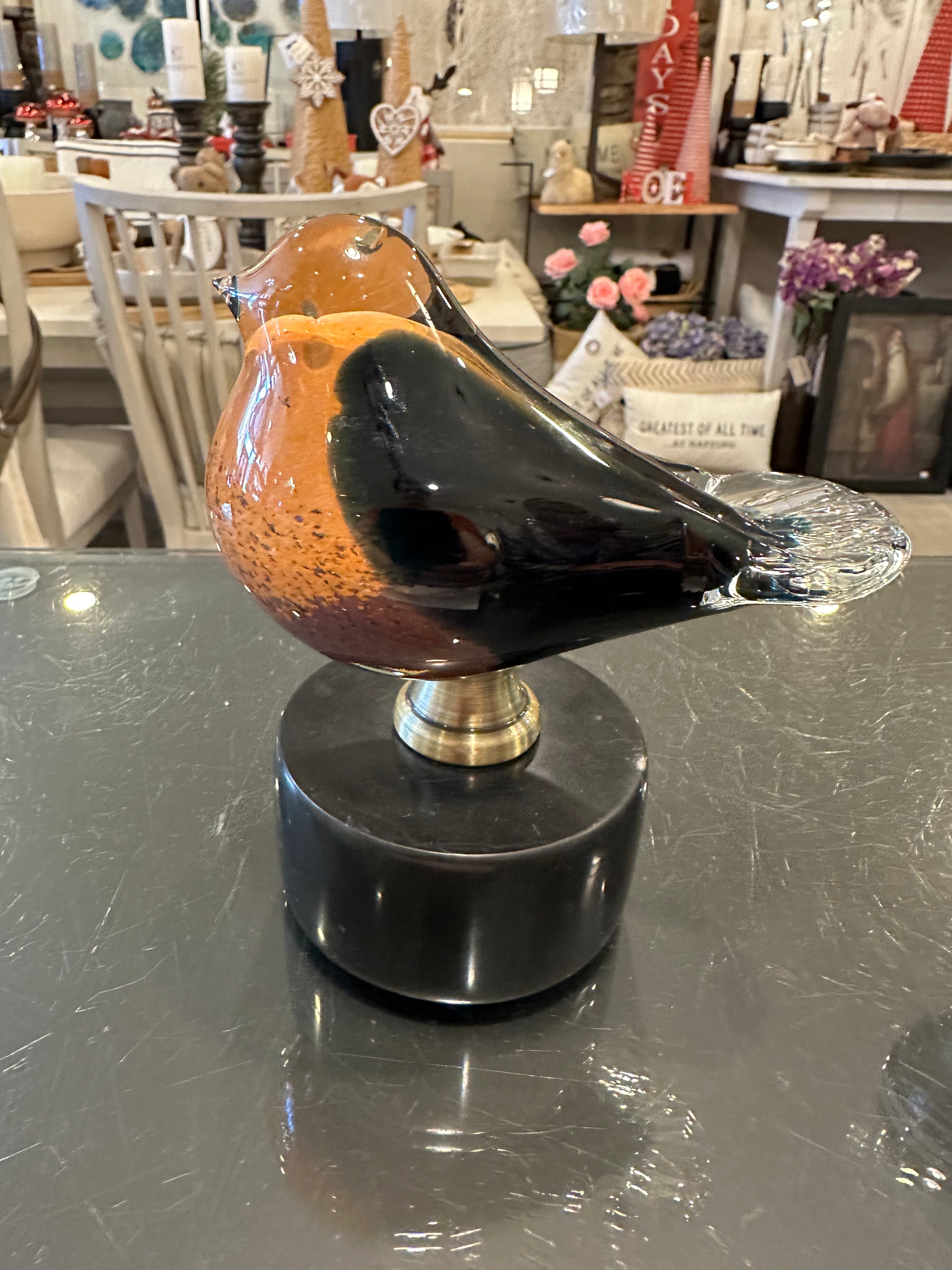 A glass figurine of a bird perched on a black marble stand, displayed on a reflective surface in a store.