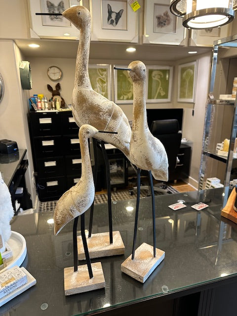 A trio of sculpted heron statues in a distressed wood finish, displayed on stands, with a framed picture in the background.