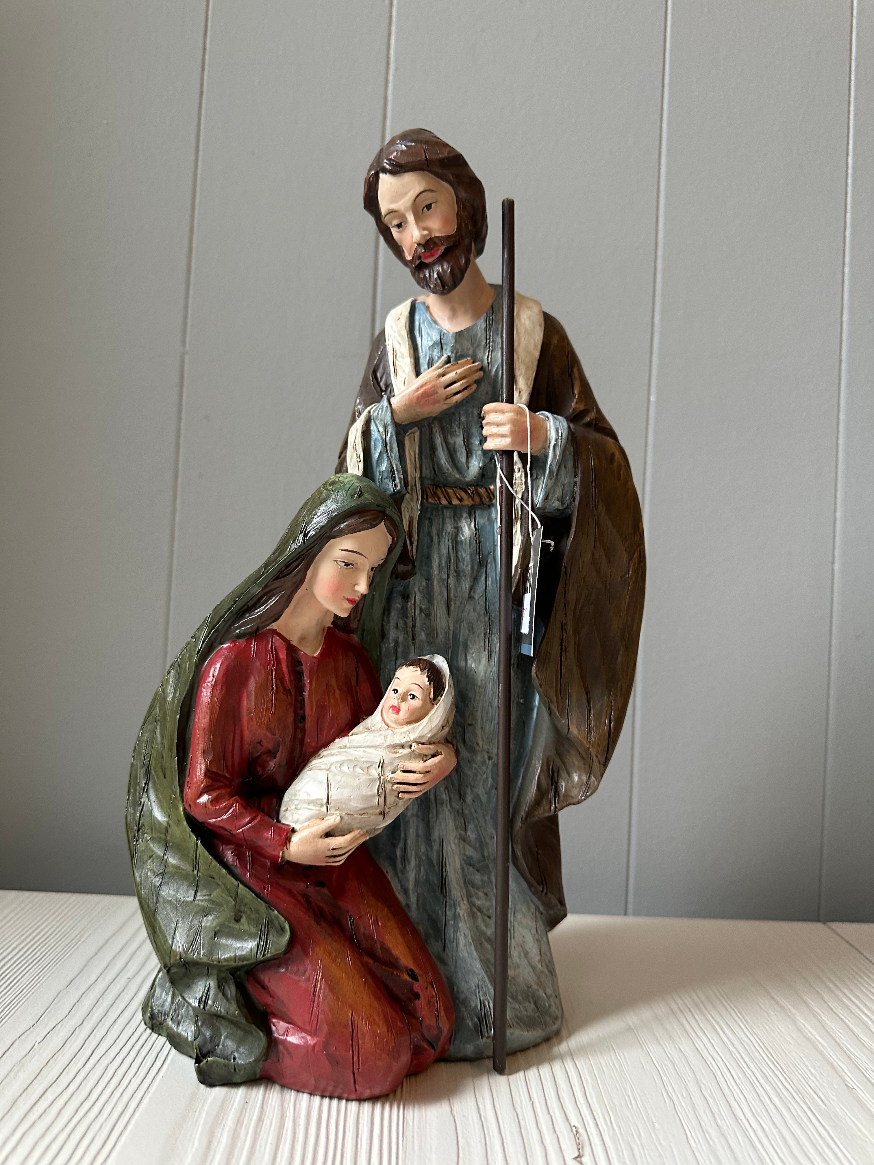 A figurine of the Holy Family, depicting Joseph in a blue robe, Mary in a red dress, and baby Jesus in her arms, held against a plain background.