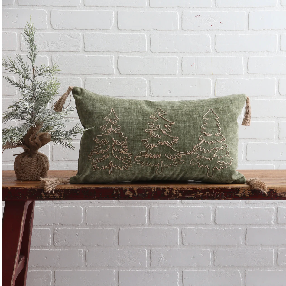 Green pillow with embroidered trees on a wooden bench against a white brick wall.