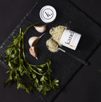 Black slate cutting board with garlic and parsley and a tipped over cainnister of Kanel Holy Grail Garlic Sale. 