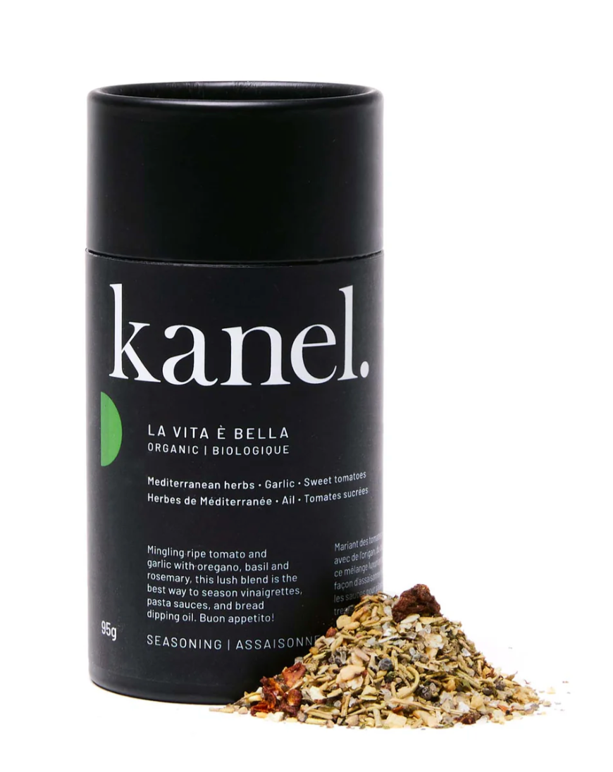 A black cylindrical container labeled 'kanel. LA VITA E BELLA' with a small pile of green herbs and tomato flakes in front of it, indicating an Italian spice blend.