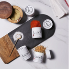 An assortment of finishing salts from the brand Kanel, displayed on a slate platter with each container labeled with different flavors such as truffle, garlic, maple, and tomato.