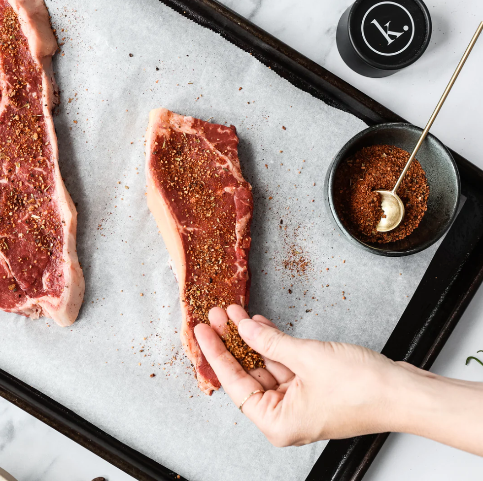 Raw steak being seasoned with spices on a baking tray.