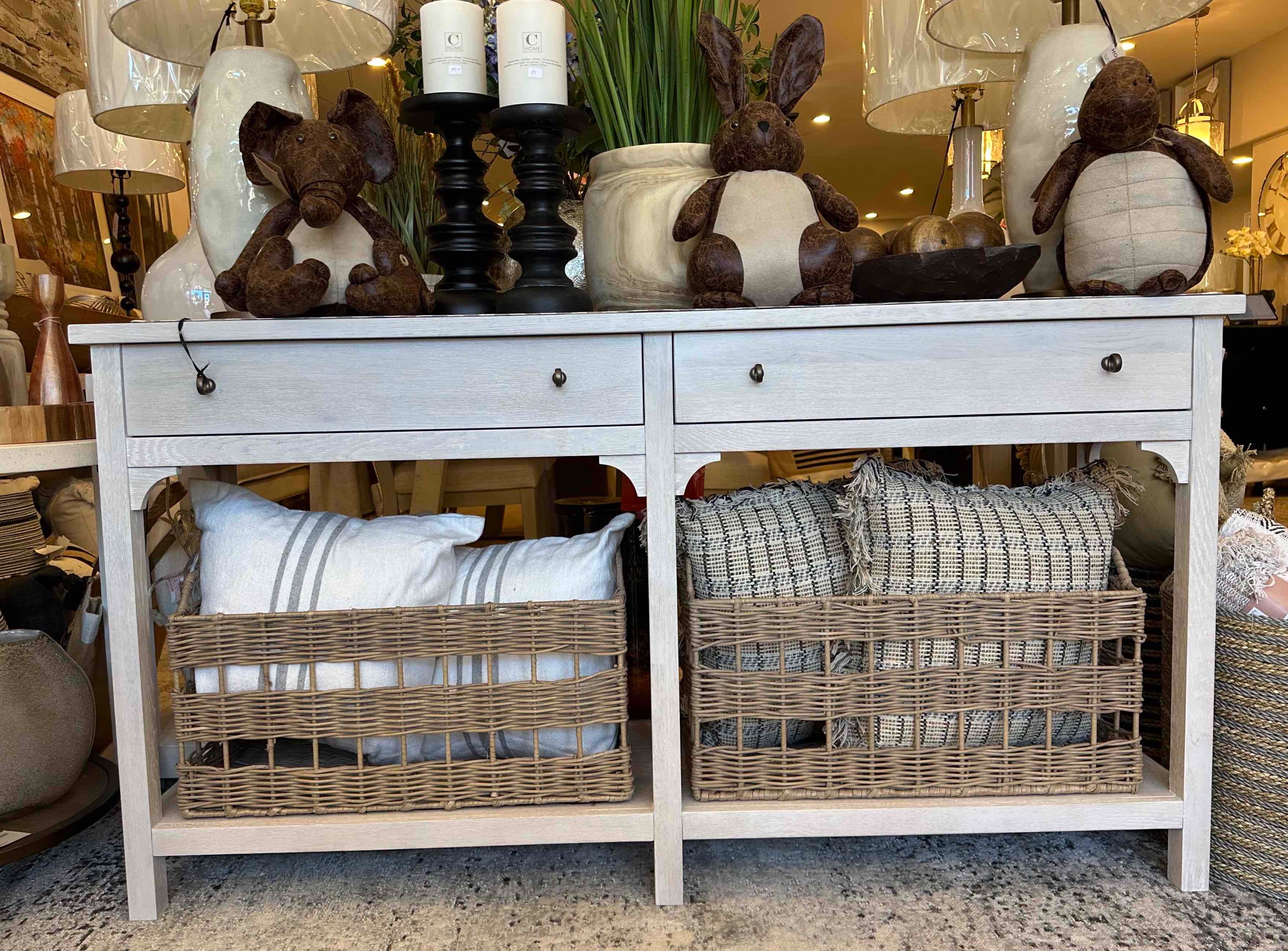 A Kinsley Console Table with two drawers and two woven baskets on a lower shelf, finished in rustic natural oak, with decorative items and pillows on top.