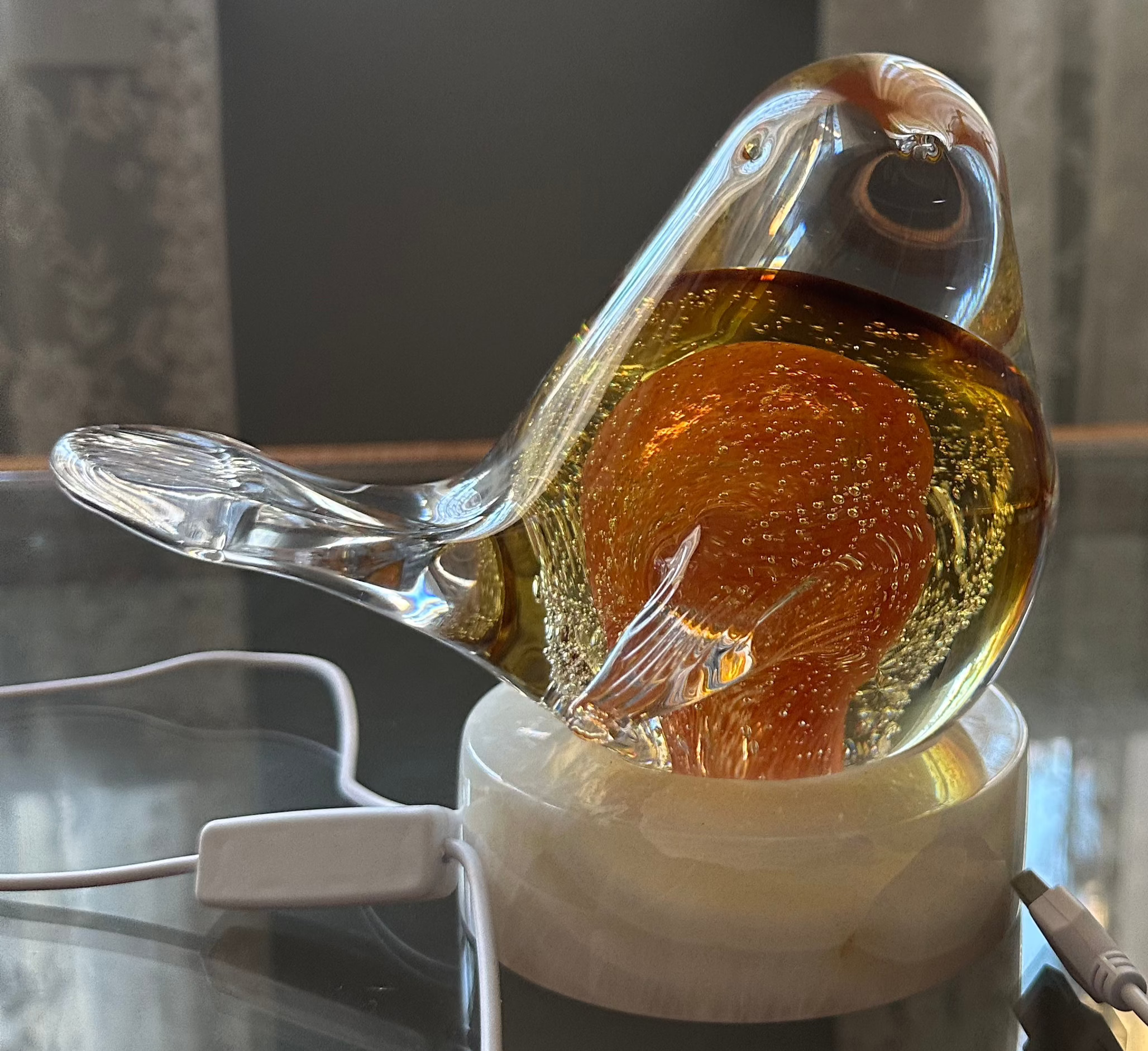 A glass bird-shaped light fixture with an orange tint, perched on a base, illuminated by a light source outside the frame.