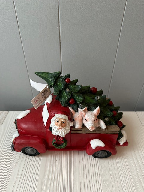 A decorative figure of a red pick-up truck with Santa Claus driving and two pink pig figures and a Christmas tree in the pick-up.