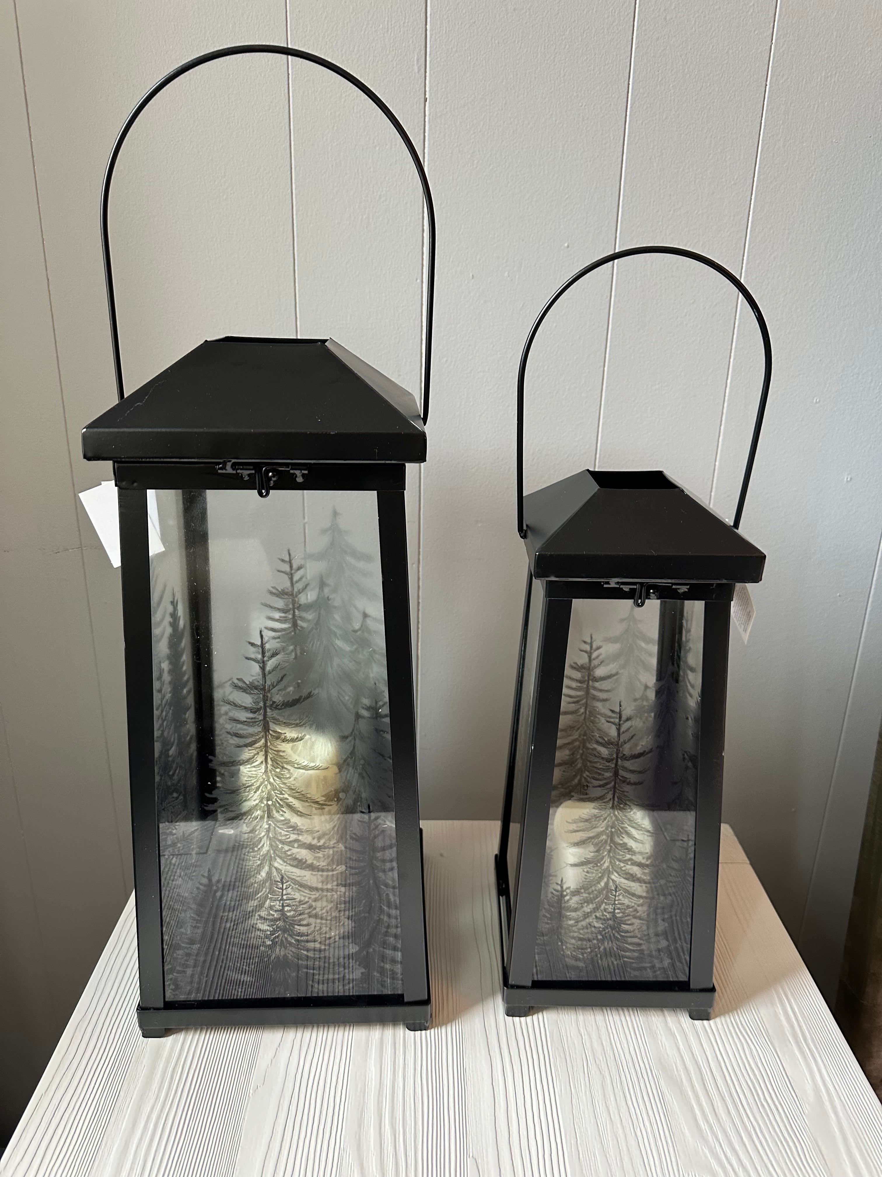 Two black LED candle lanterns with glass panels that have trees.