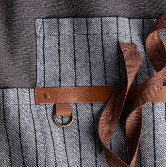 Close up of the Let's Get Grillin' Apron showing grey material with thin black pinstripes faux brown leather detailing and a brown tie. 