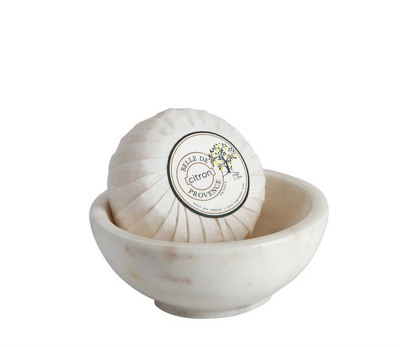 A round soap wrapped in white paper with a label that reads "belle De Provence Citron" in a small marble bowl. 