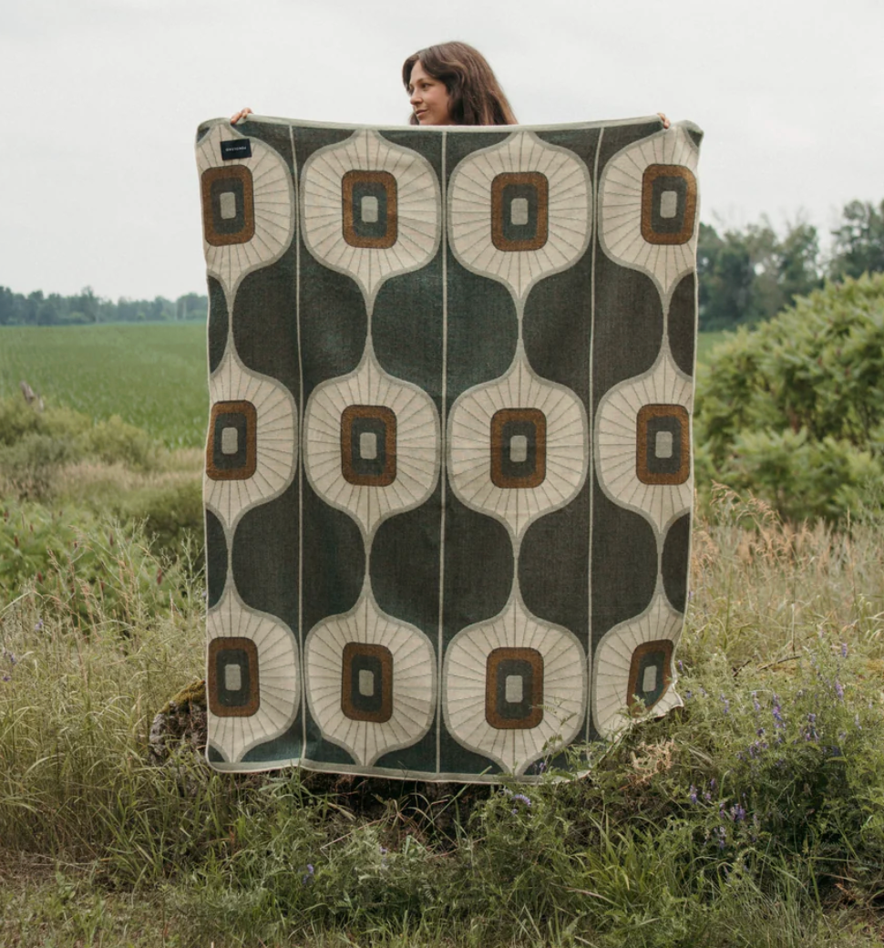 Person holding a patterned green, cream and brown throw blanket in a grassy field