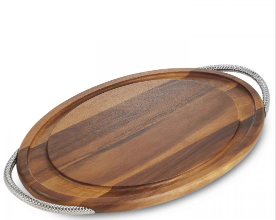 An oval-shaped carving board made of acacia wood with a chrome braid handle.