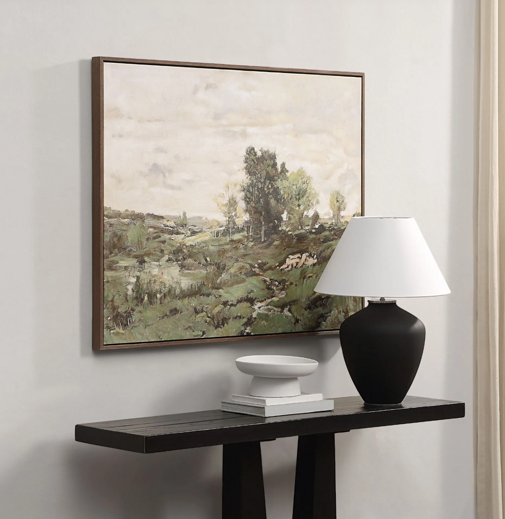 Framed landscape painting on a wall above a console table with a lamp and decorative items.