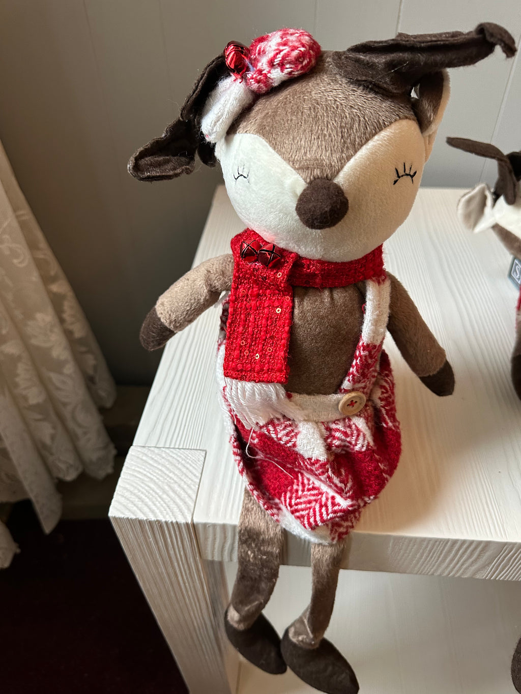 A plush toy resembling a sitting reindeer with a red and white checkered dress and a brown bow on its head, placed on a wooden surface.