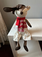 A plush toy resembling a sitting reindeer with a red and white checkered shirt, white shorts and a red scarf on a wooden surface