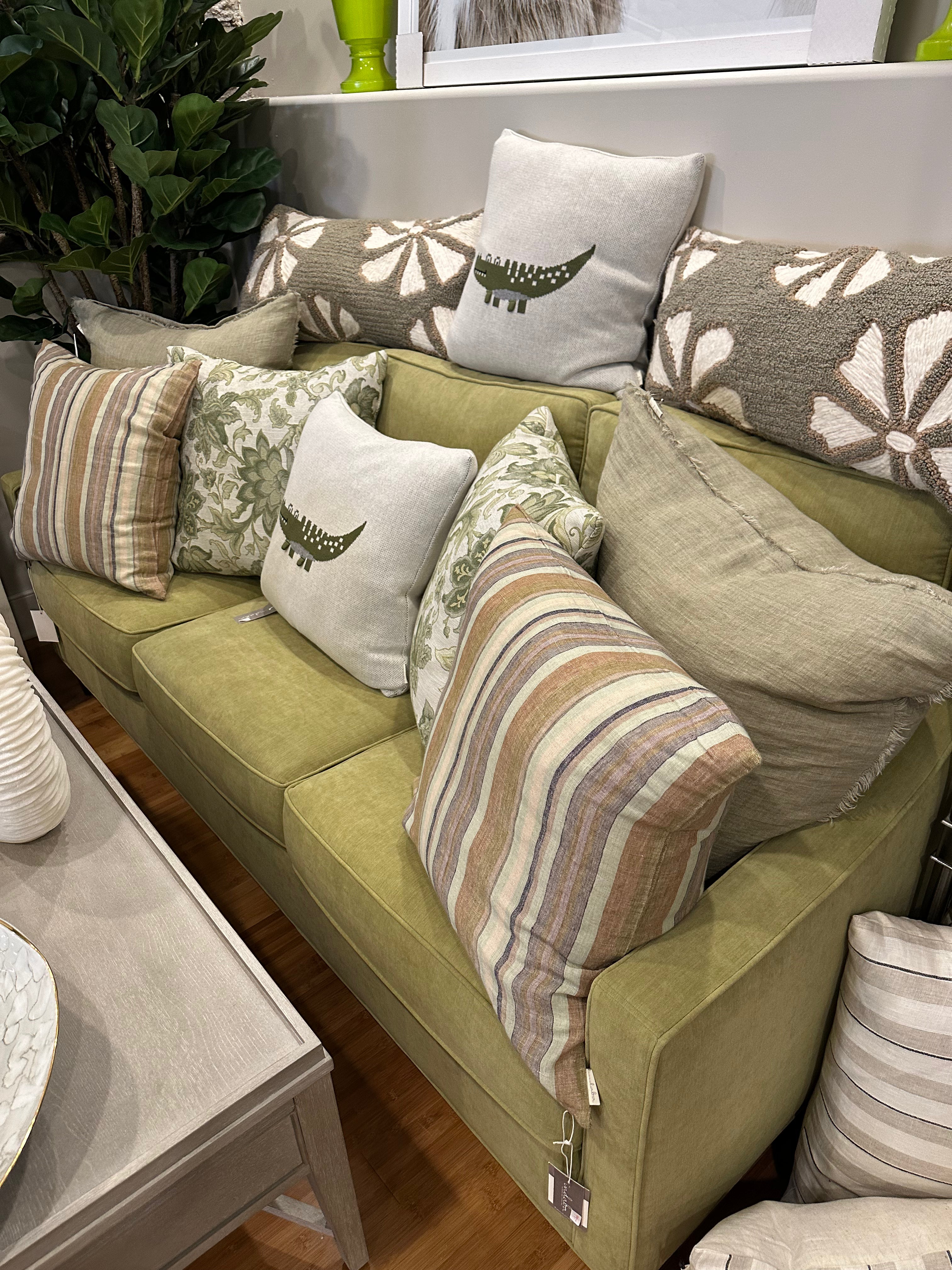 A green sofa with various styled pillows arranged on it, displayed in a room setting with a plant in the background.