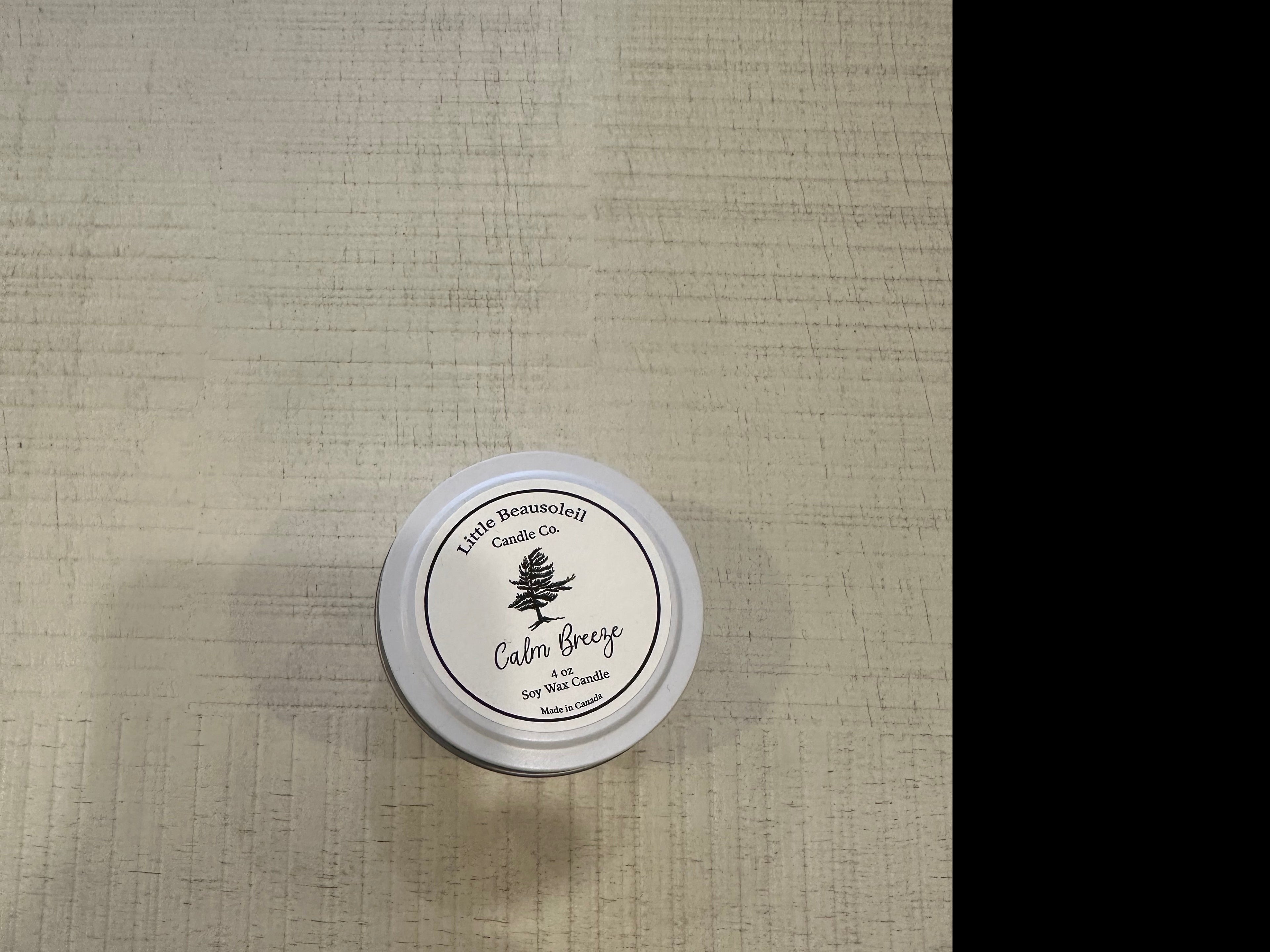 A tin candle with a label that reads 'Little Beausoleil - Calm Breeze' placed on a textured surface.