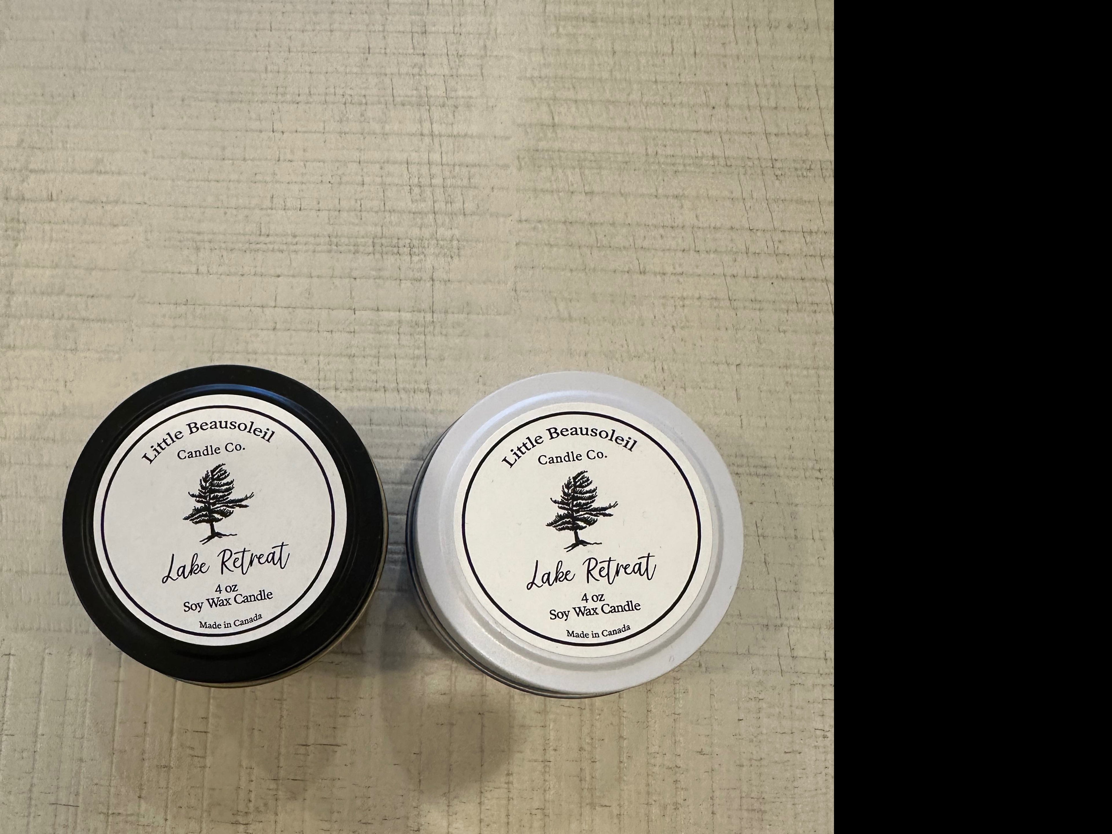 Two tin candles in black jars with white labels, one labeled 'Lake Retreat' and the other with a tree logo, presumably the scent wick candle.