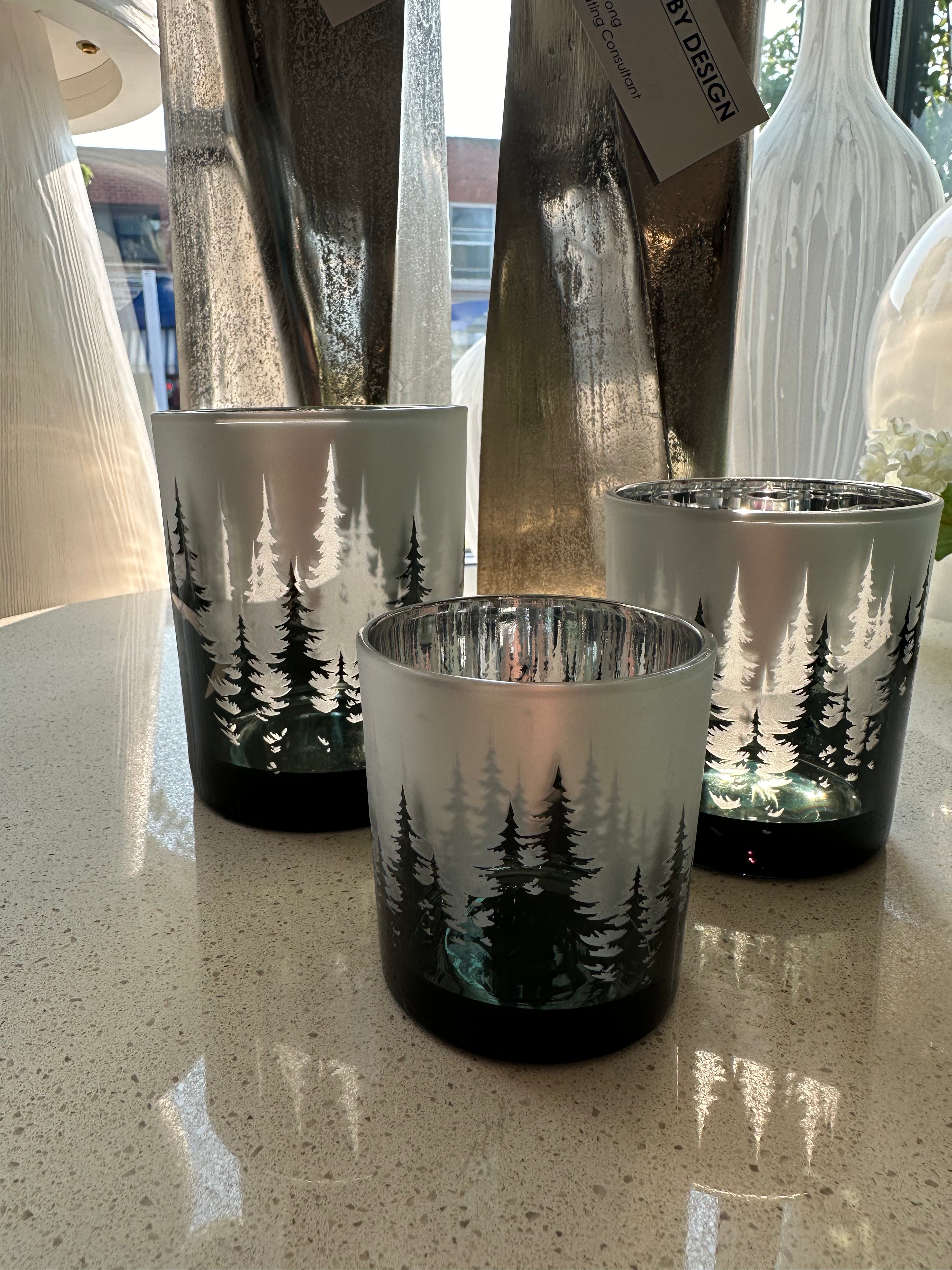 A set of three glass candle holders with a forest and trees design, displayed on a table.